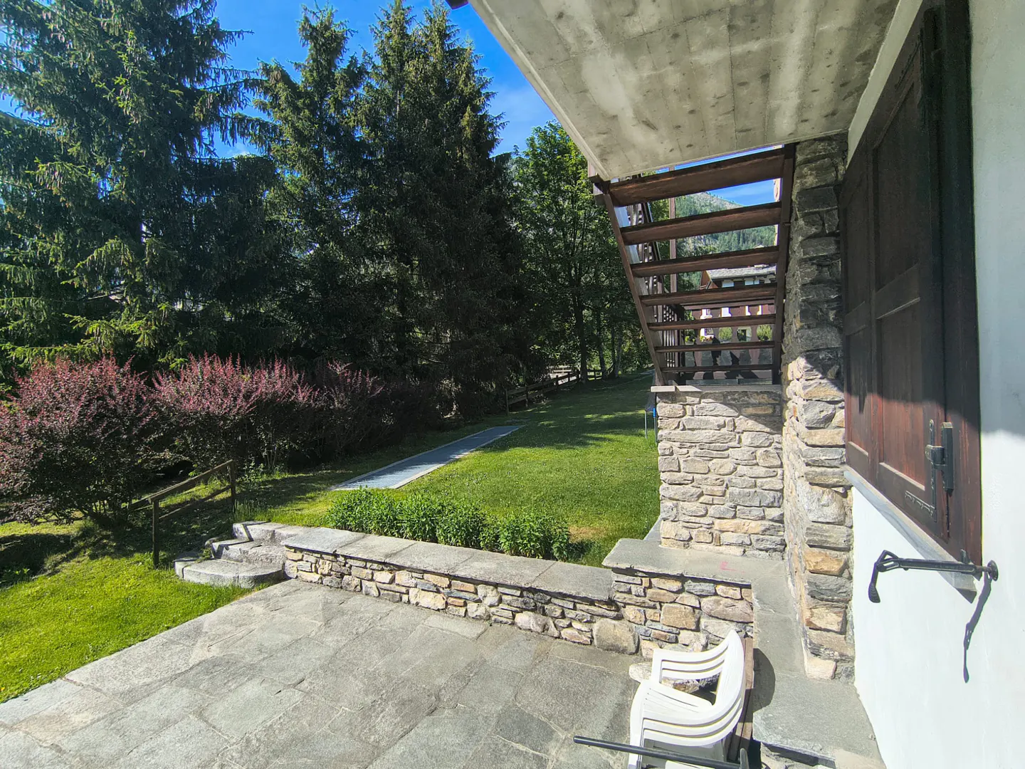 Stone patio with a low wall, white plastic chair, and wooden stairs leading to a lush green yard with tall pine trees.