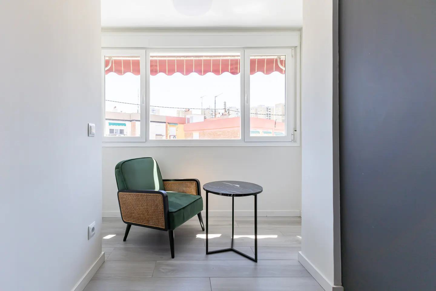 Bright room with a green armchair, wicker accents, and a black marble side table near a window with a red awning.