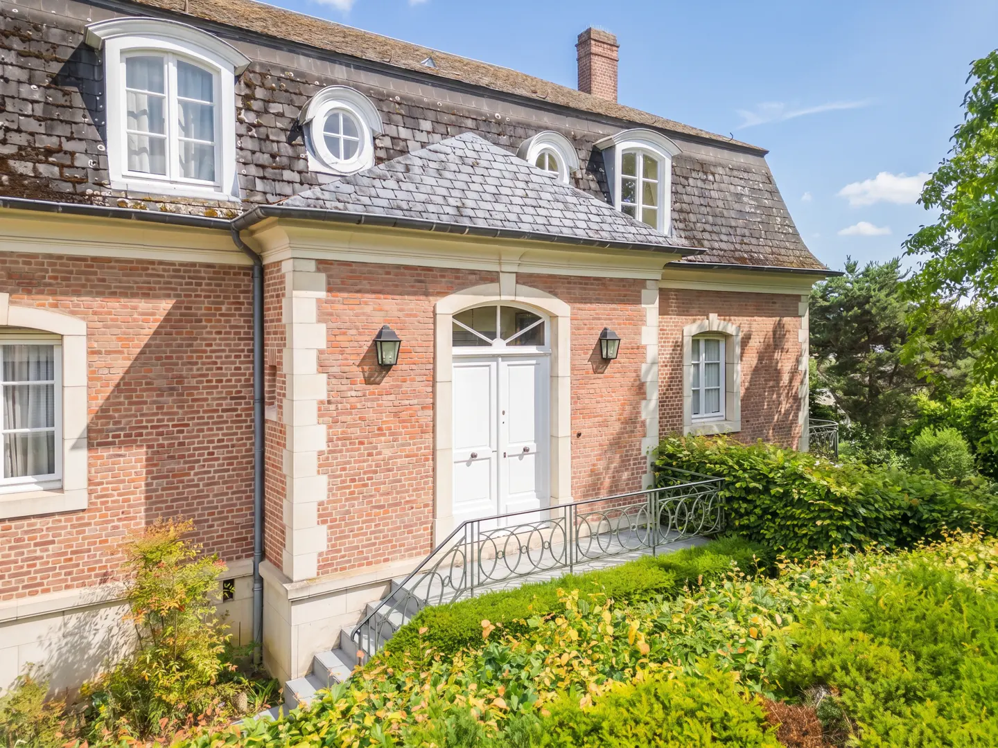 A red brick house with a slate roof, white double doors, and a metal railing leading up to the entrance.