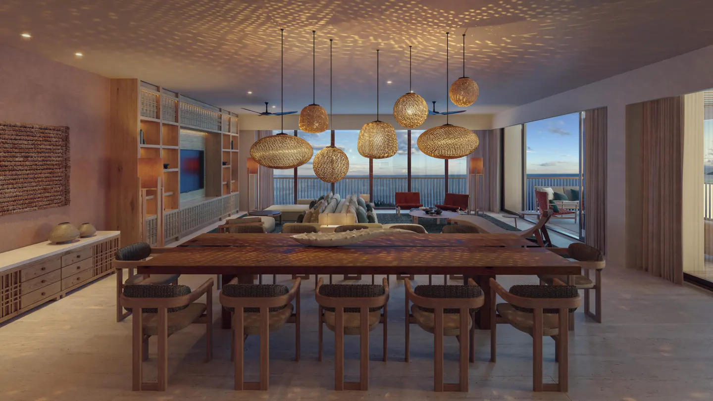 A spacious dining room with a long wooden table and chairs, lit by woven pendant lights. Large windows offer ocean views.