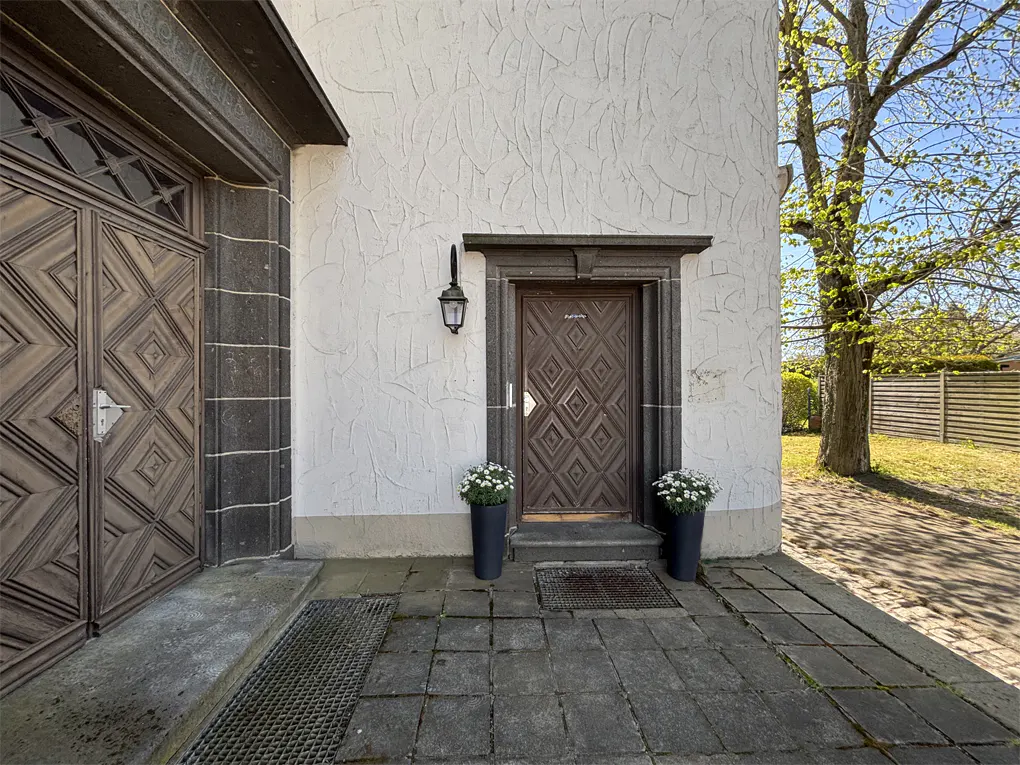 Exterior view of a building with a textured white wall, a brown diamond-patterned door, and potted white flowers on either side.