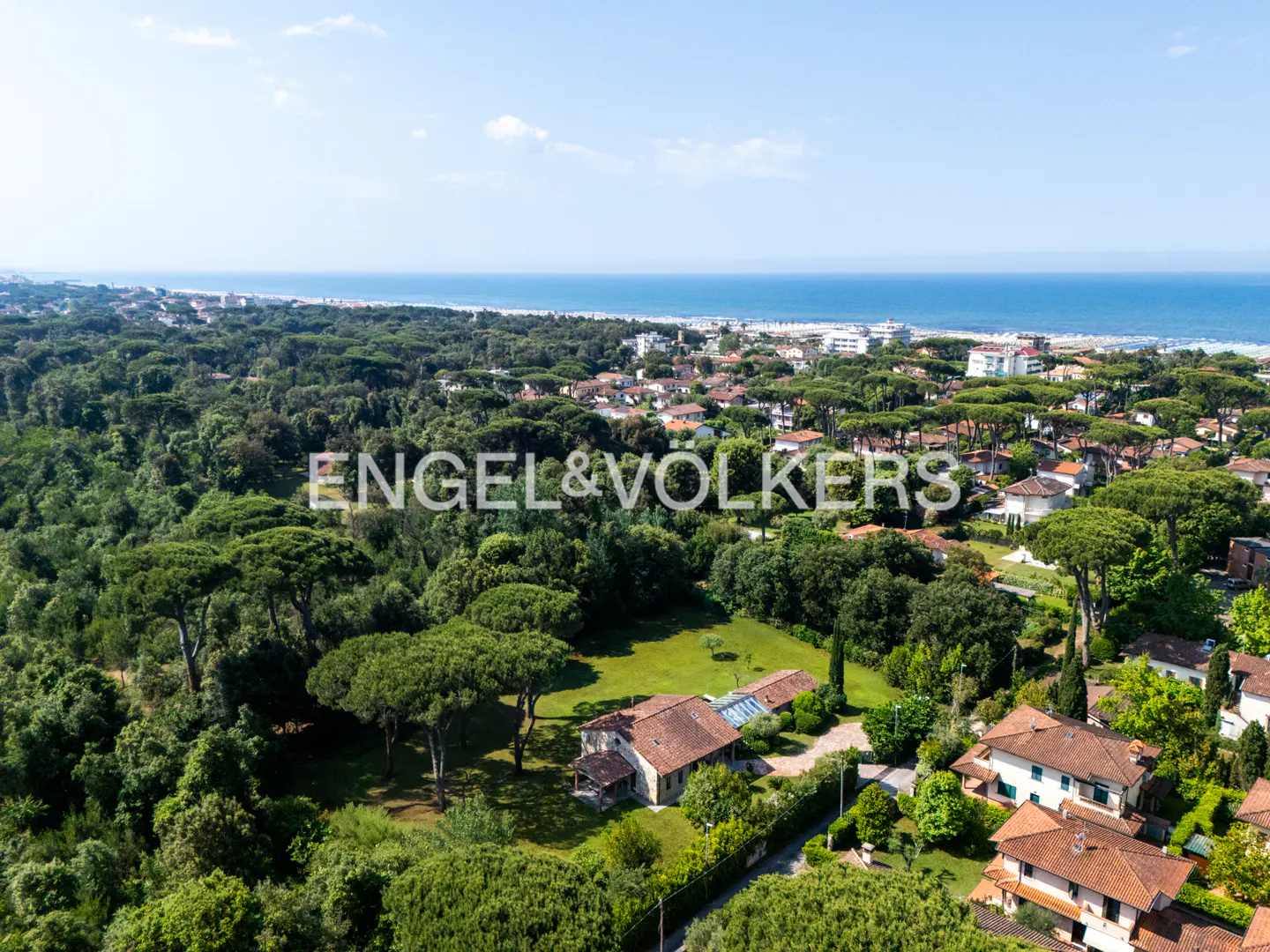 Aerial view of a villa with a terracotta roof nestled in a lush green forest near the Mediterranean coastline.