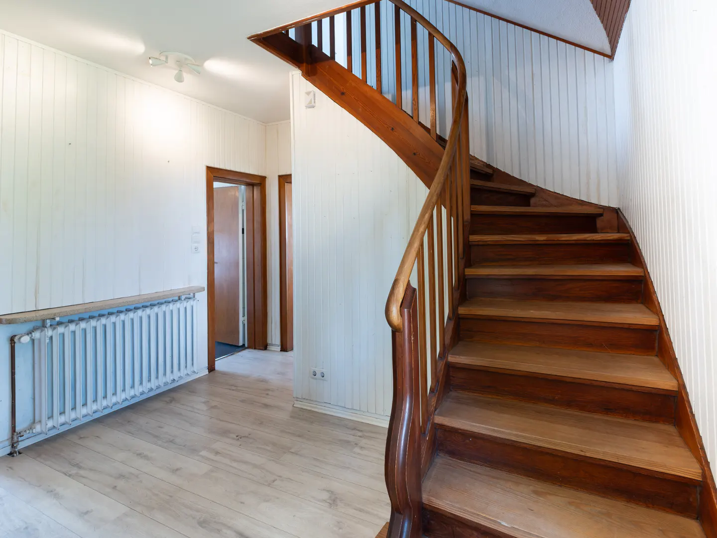 Curved wooden staircase with a railing in a hallway with white paneled walls, light wood floors, and a white radiator.