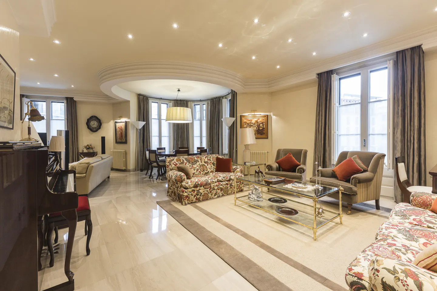 Bright, spacious living room with marble floors, floral sofas, armchairs, and a grand piano. Large windows with gray curtains.
