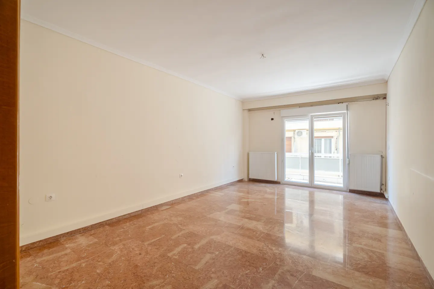 Empty room with cream walls, marble floors, and a sliding glass door to a balcony. Two white radiators are visible.