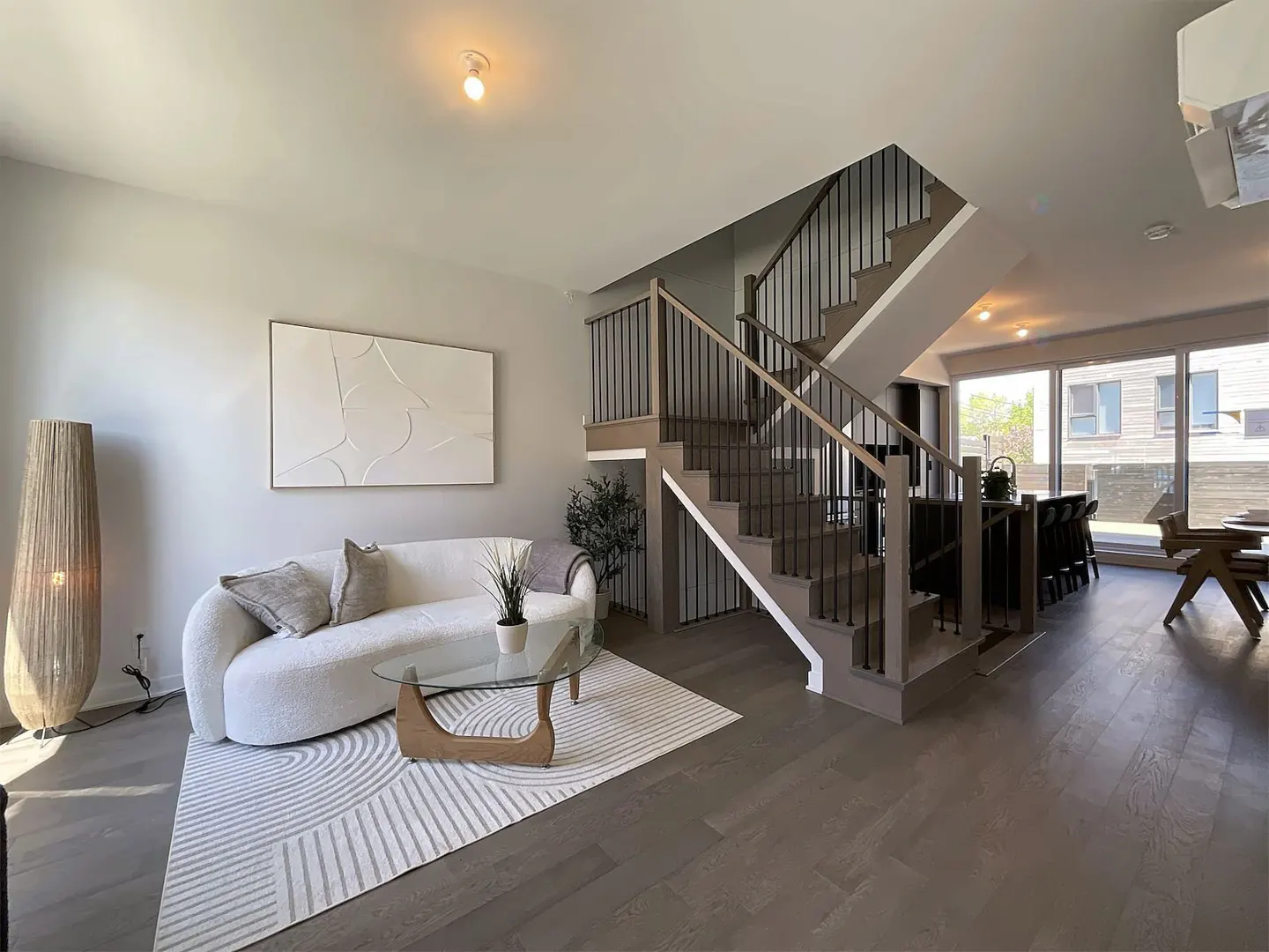 Open-concept living room with a white sofa, glass coffee table, and a staircase with black railings. Hardwood floors and a large abstract art piece.