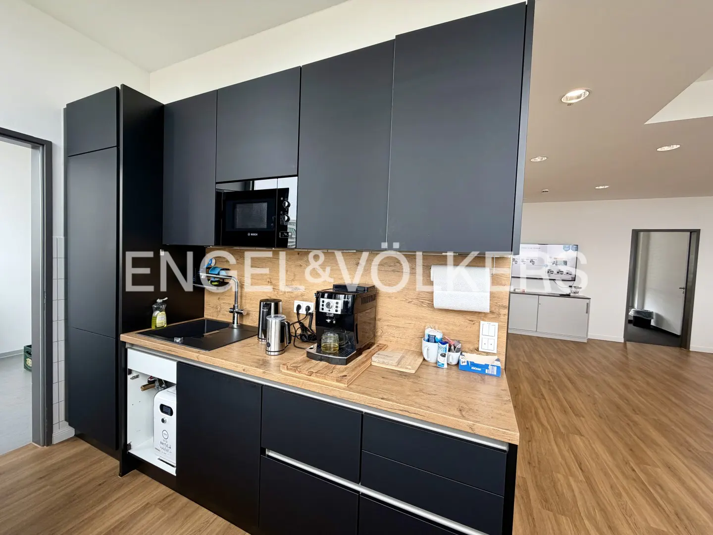 A modern kitchen with black cabinets, wood countertop, microwave, coffee maker, and sink. The floor is wood and there is an open doorway in the background.