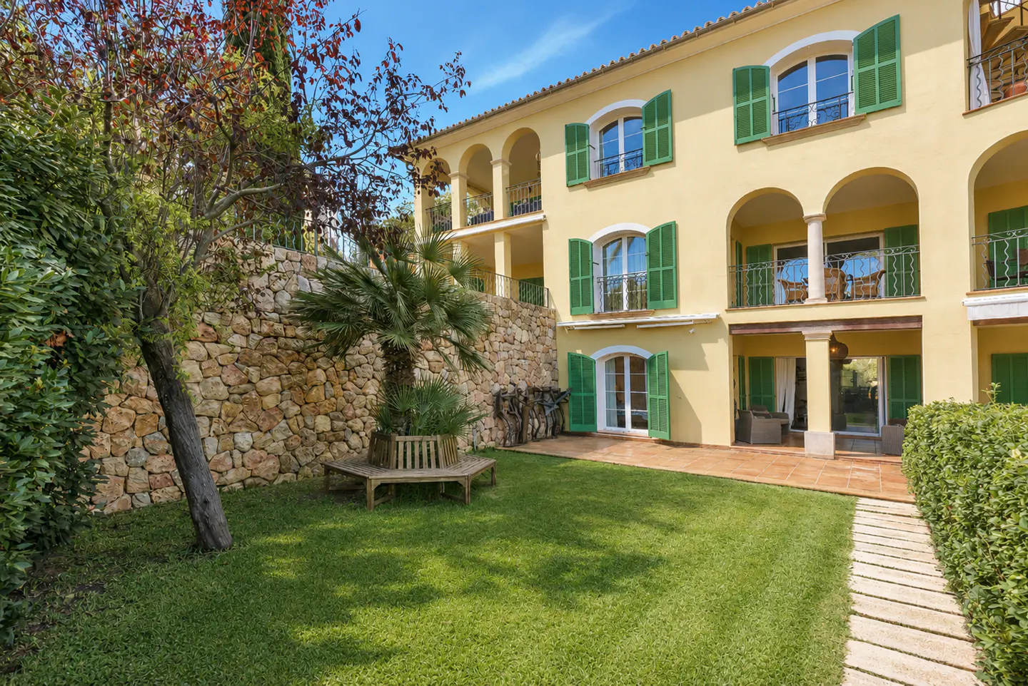 Two-story yellow house with green shutters, stone wall, and green lawn. A palm tree and bench sit in the yard.