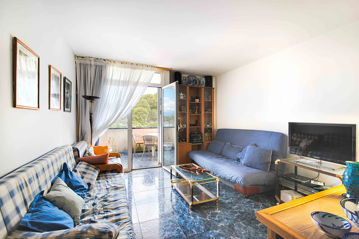 Bright living room with blue tile floor, sofas, TV, and balcony with outdoor seating.