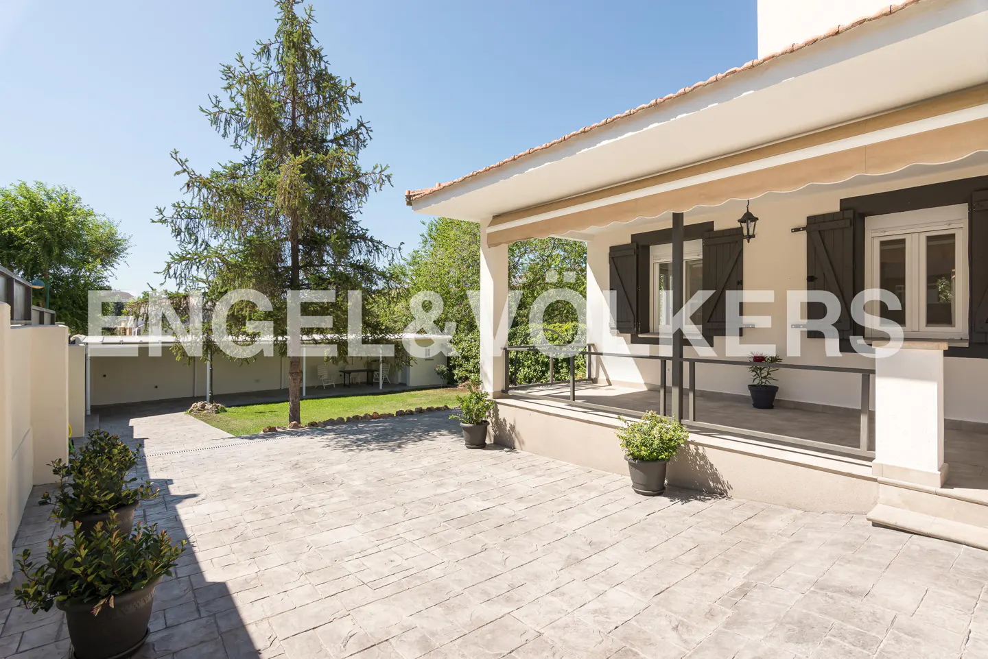 Exterior view of a cream-colored house with dark brown shutters and a stone patio. A tree and greenery are visible in the background.