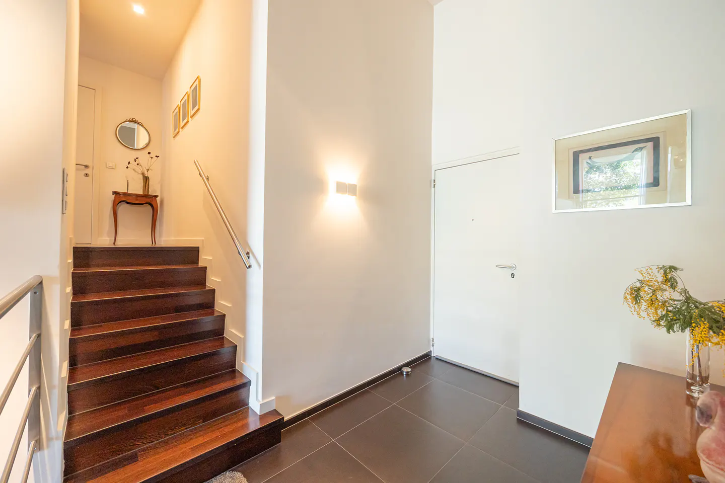 Interior view of a home's entryway with dark wood stairs, white walls, and dark tile floors. A white door and framed art are visible.