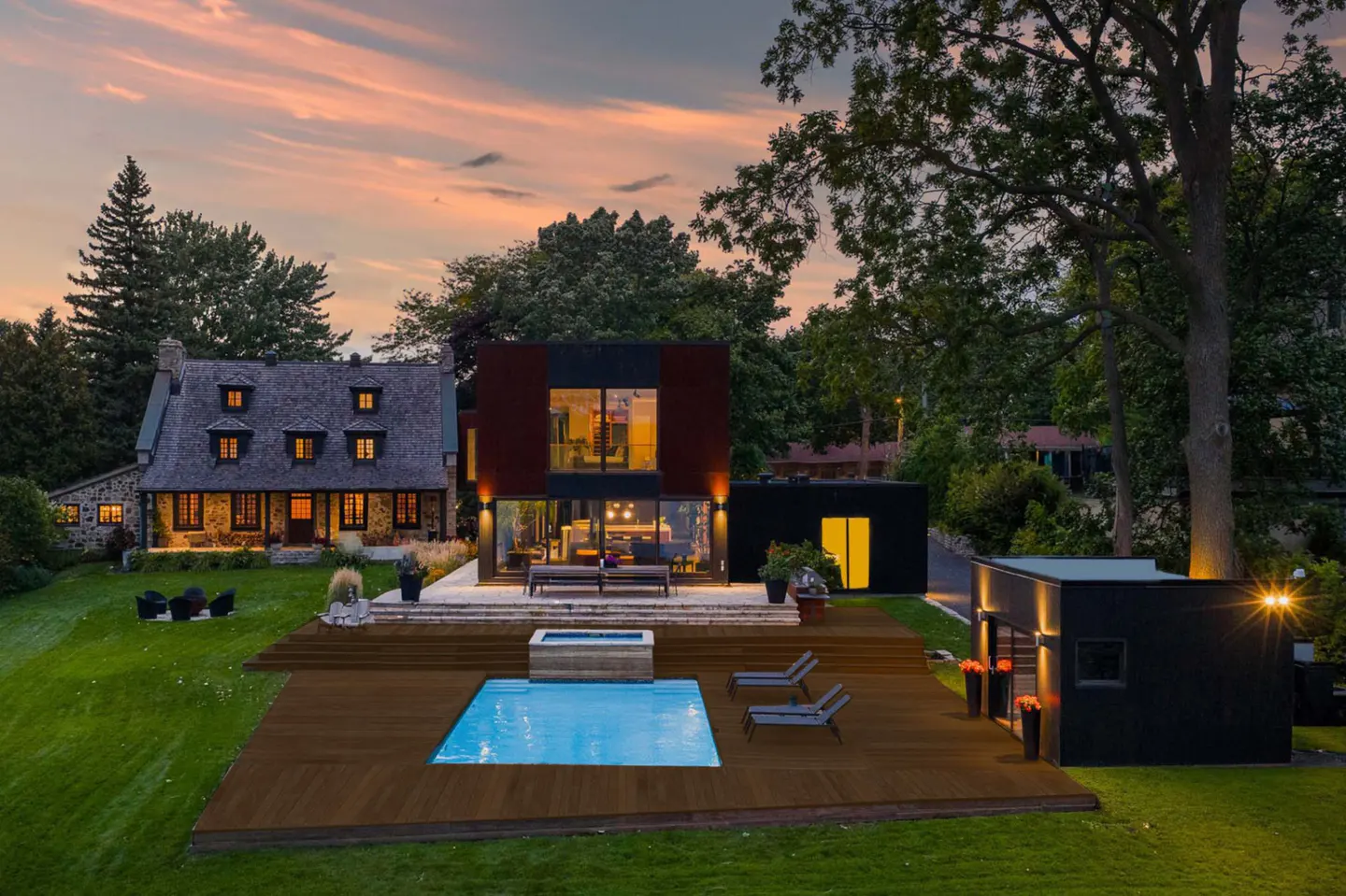 Exterior view of a modern home with a pool and deck at dusk, next to a traditional stone house.