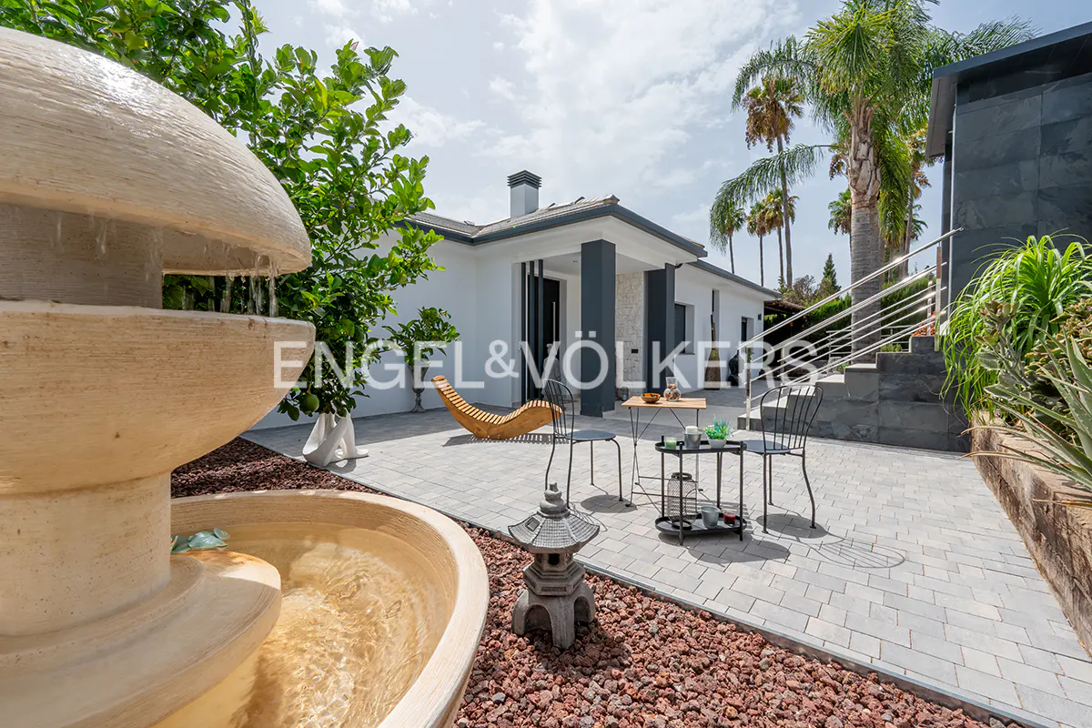 Outdoor patio with a fountain, table, chairs, and a modern white house with dark gray pillars and trim. Palm trees are in the background.