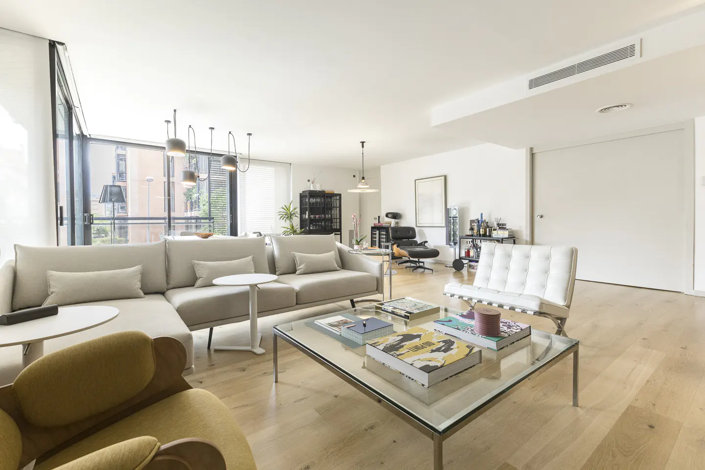 Bright, modern living room with a beige sectional, white leather chair, glass coffee table, and light wood flooring.