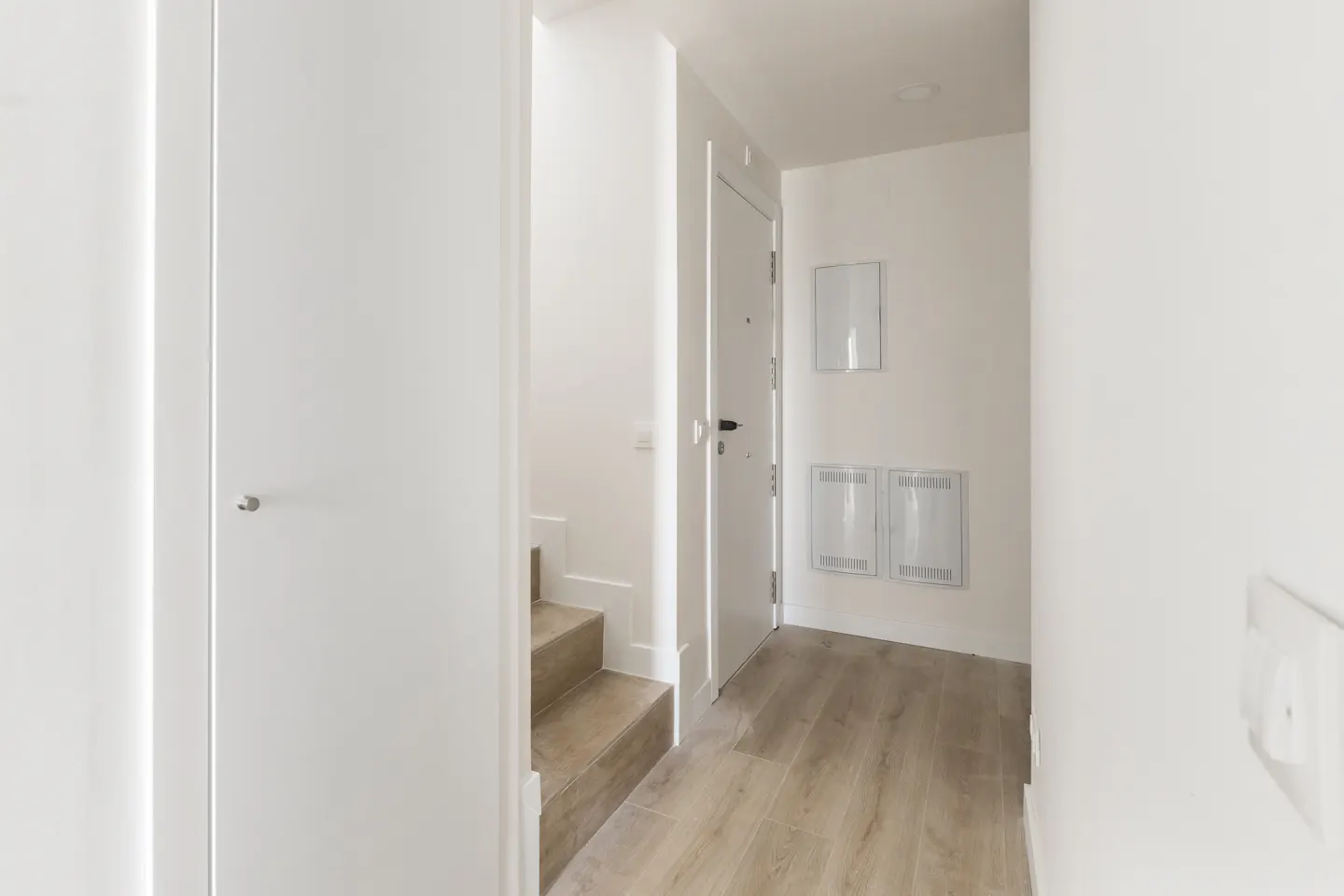 Bright hallway with white walls, light wood flooring, a white door, and wooden stairs leading to an upper level.