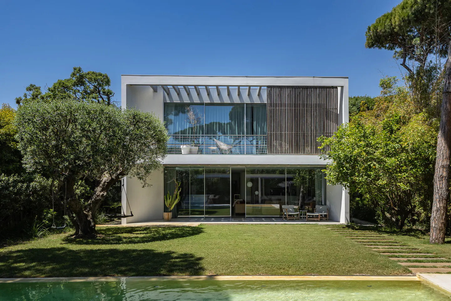 Modern two-story house with a pool, lawn, and trees. The house has large glass windows and a balcony with a hammock.