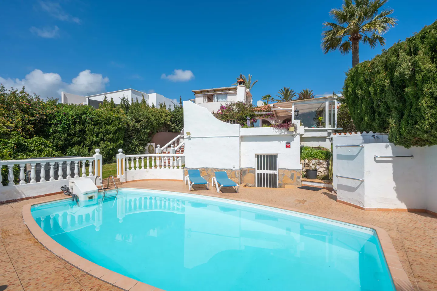 A bright, sunny day highlights a turquoise pool with two blue lounge chairs nearby. White buildings and lush greenery surround the pool area.