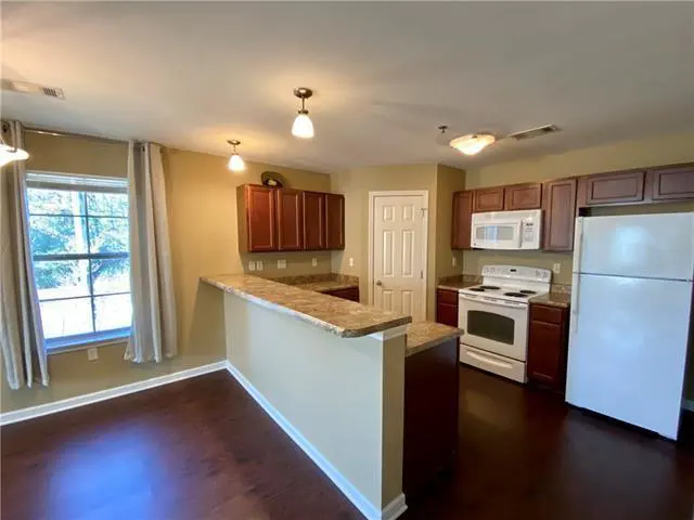 A kitchen with dark wood floors, brown cabinets, white appliances, and a breakfast bar. A window with curtains is on the left.