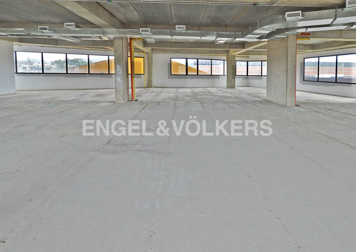 An empty, unfinished office space with concrete floors, support pillars, and large windows.