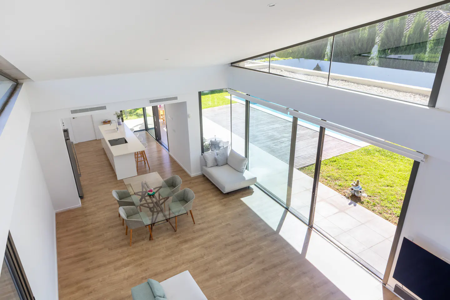 Bright, modern open-plan living space with wood floors, glass dining table, white kitchen island, and large windows overlooking a pool and green lawn.