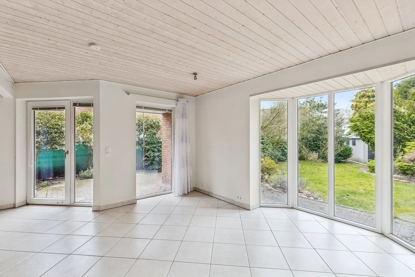 Bright sunroom with white tile floor, wood ceiling, and large windows overlooking a green lawn and garden.