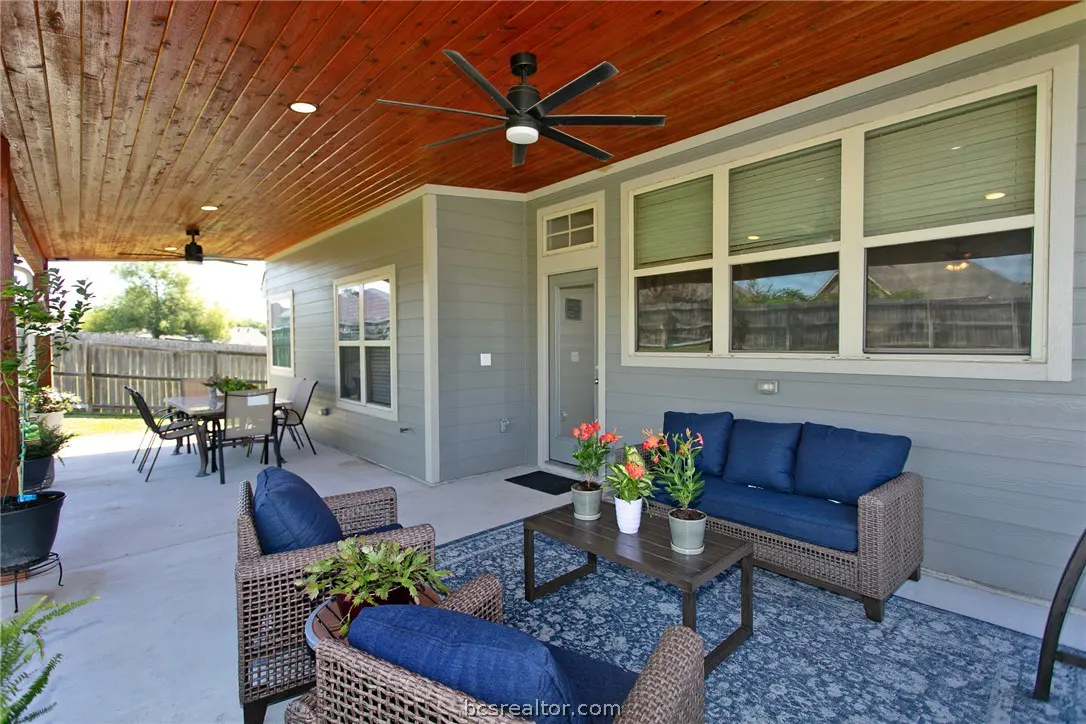 Covered patio with wood ceiling, ceiling fan, and gray siding. Wicker furniture with blue cushions sits on a blue rug. Table and chairs in the background.