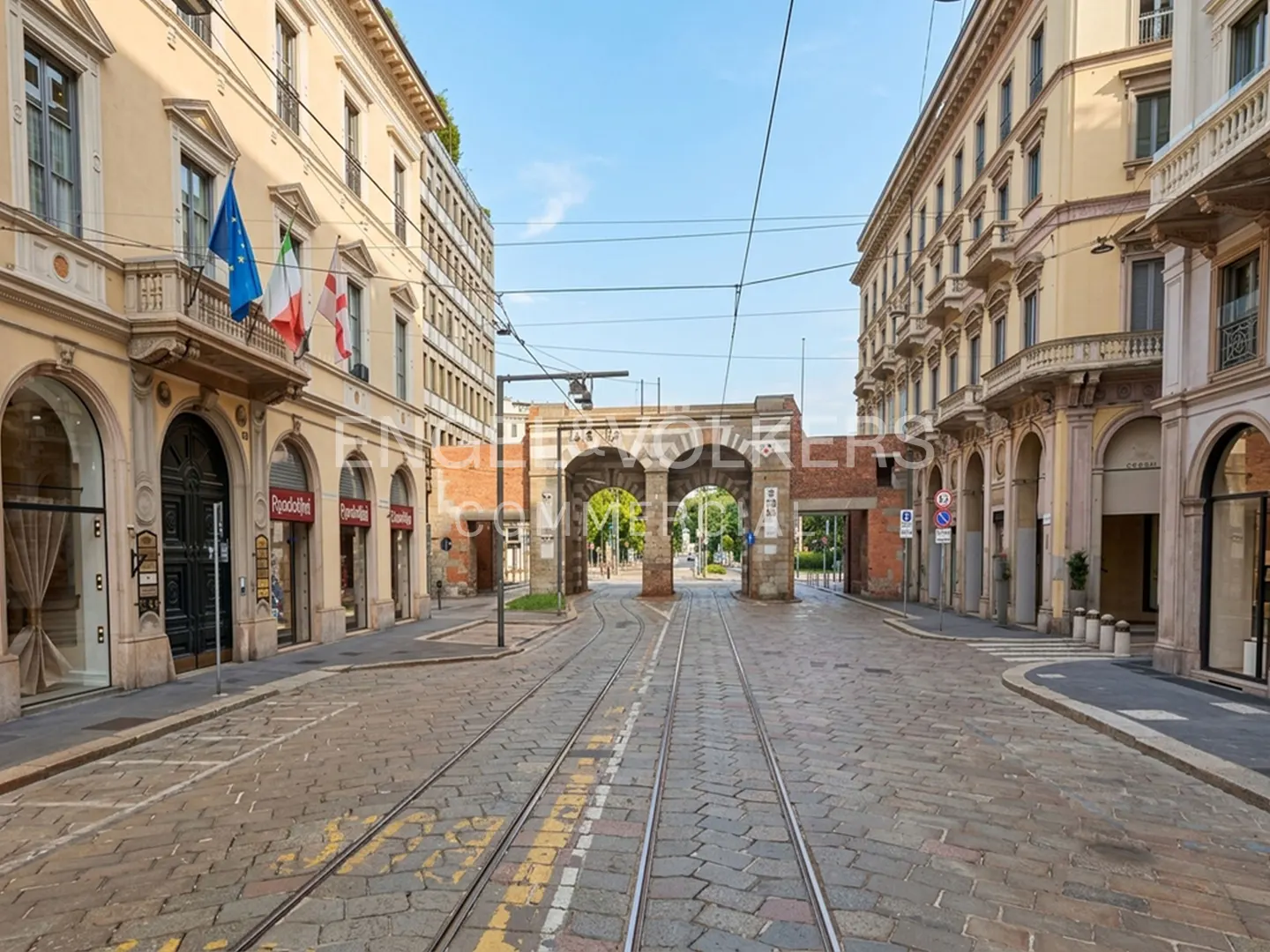 Street view of Milan, Italy, with tram tracks leading to an archway. Buildings line the street, with flags on one side.