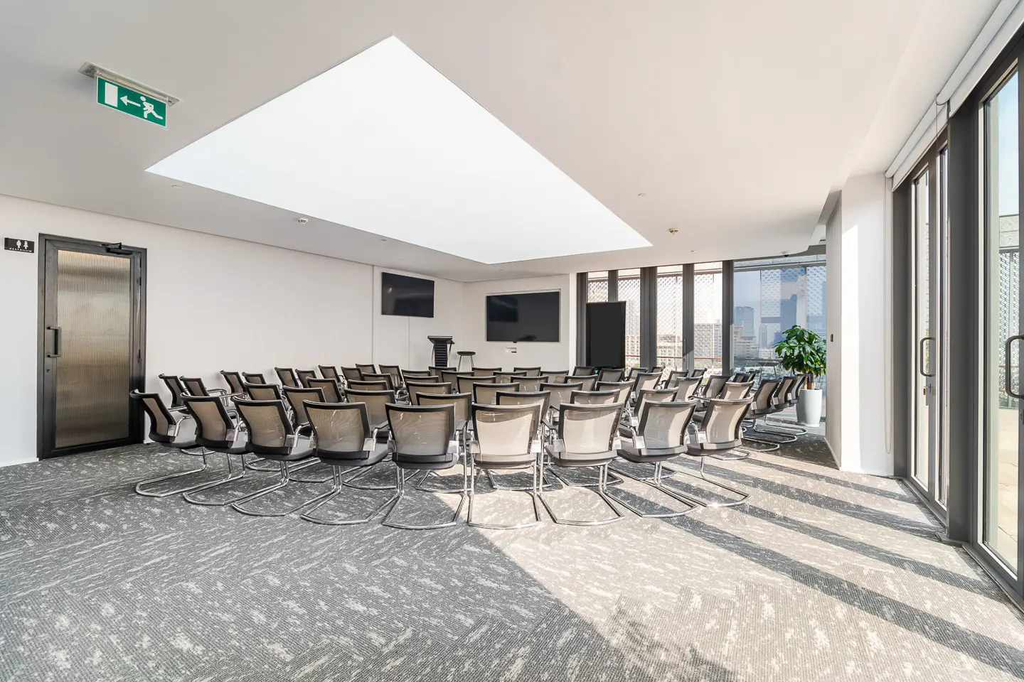 Bright conference room with rows of gray chairs facing a stage with two screens. Sunlight streams through large windows.