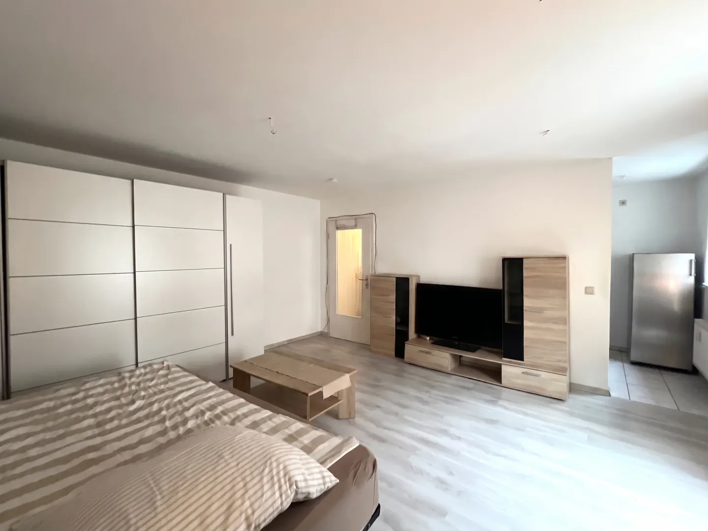 A bedroom with a large white wardrobe, a striped bed, a wooden coffee table, a TV unit, and a stainless steel refrigerator.