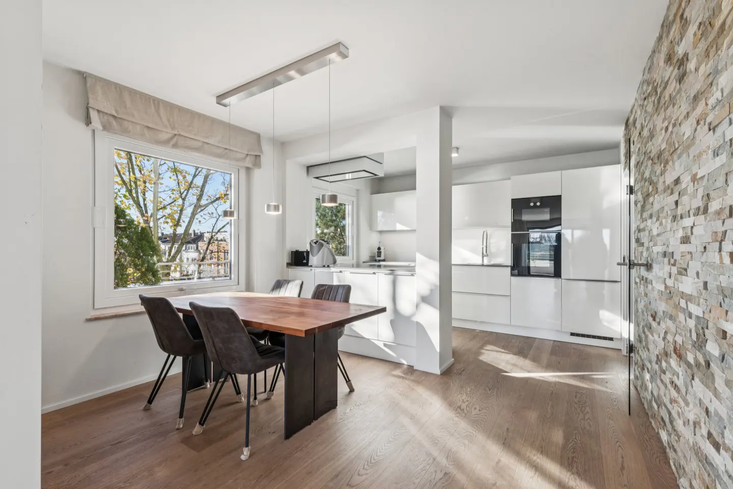 Bright, modern kitchen with wood floors, a dining table with four chairs, and white cabinets. A stone accent wall adds texture.
