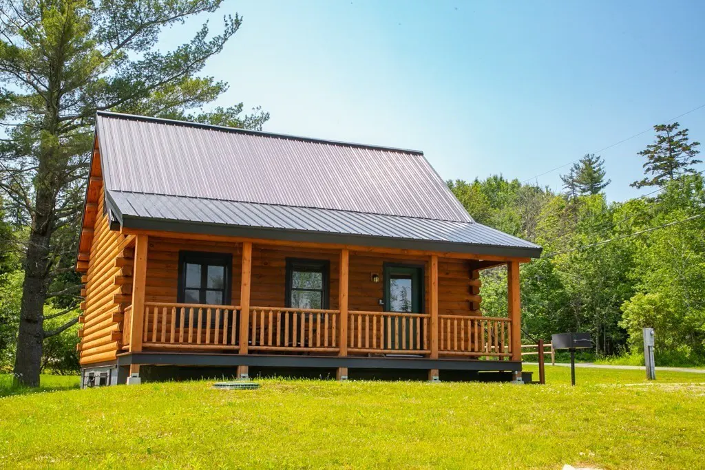 A cozy log cabin with a metal roof and porch, set on a green lawn with trees in the background.