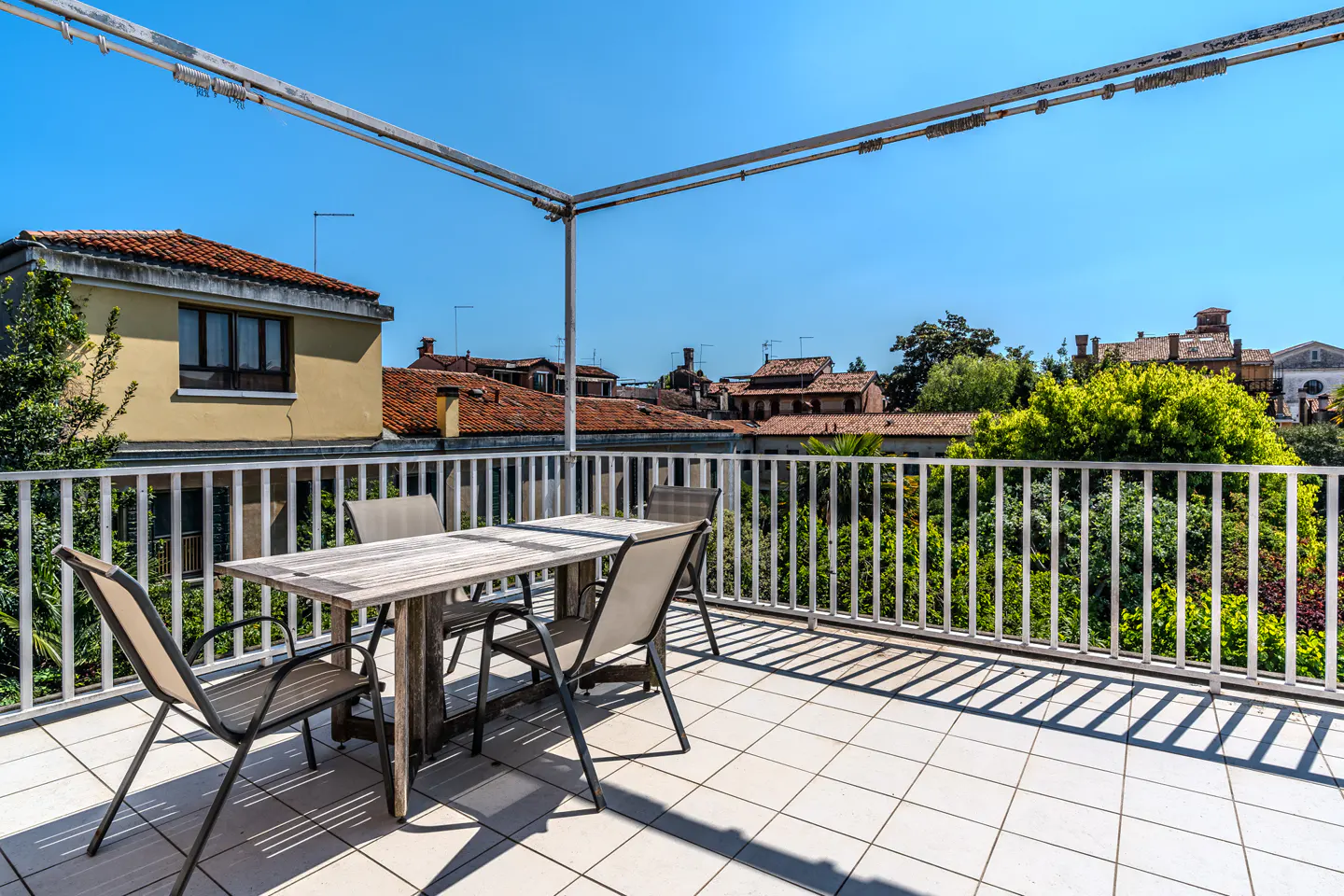 Outdoor patio with a wooden table and chairs on a tiled floor, surrounded by a white railing, with a view of rooftops and trees.