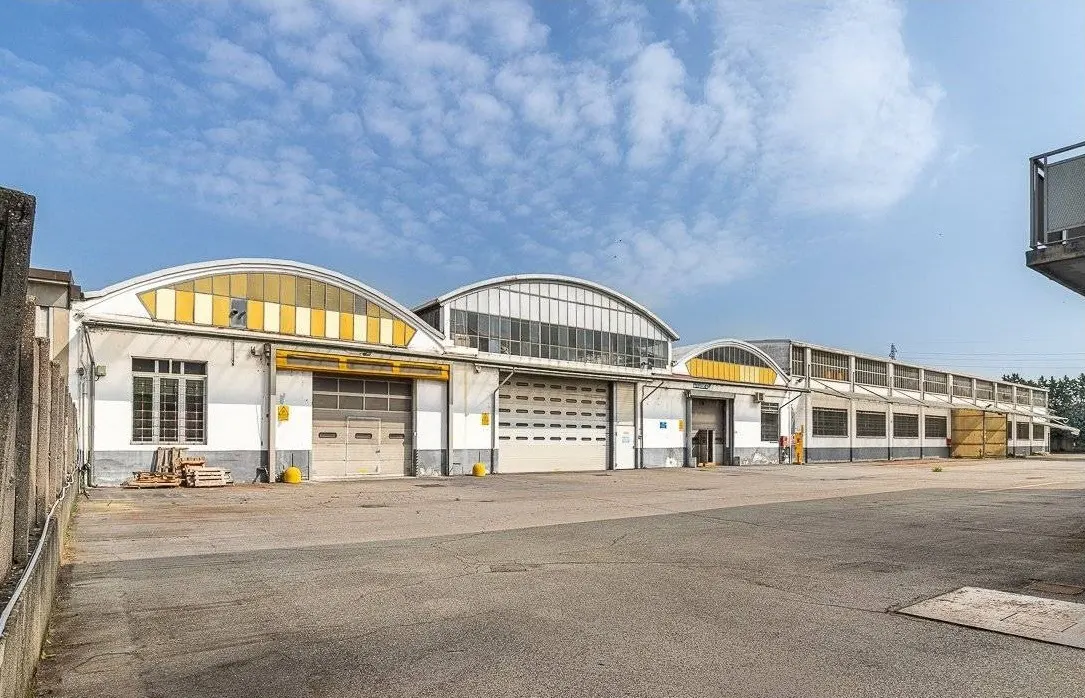 Exterior of a white industrial building with multiple garage doors and arched windows under a blue, cloudy sky.