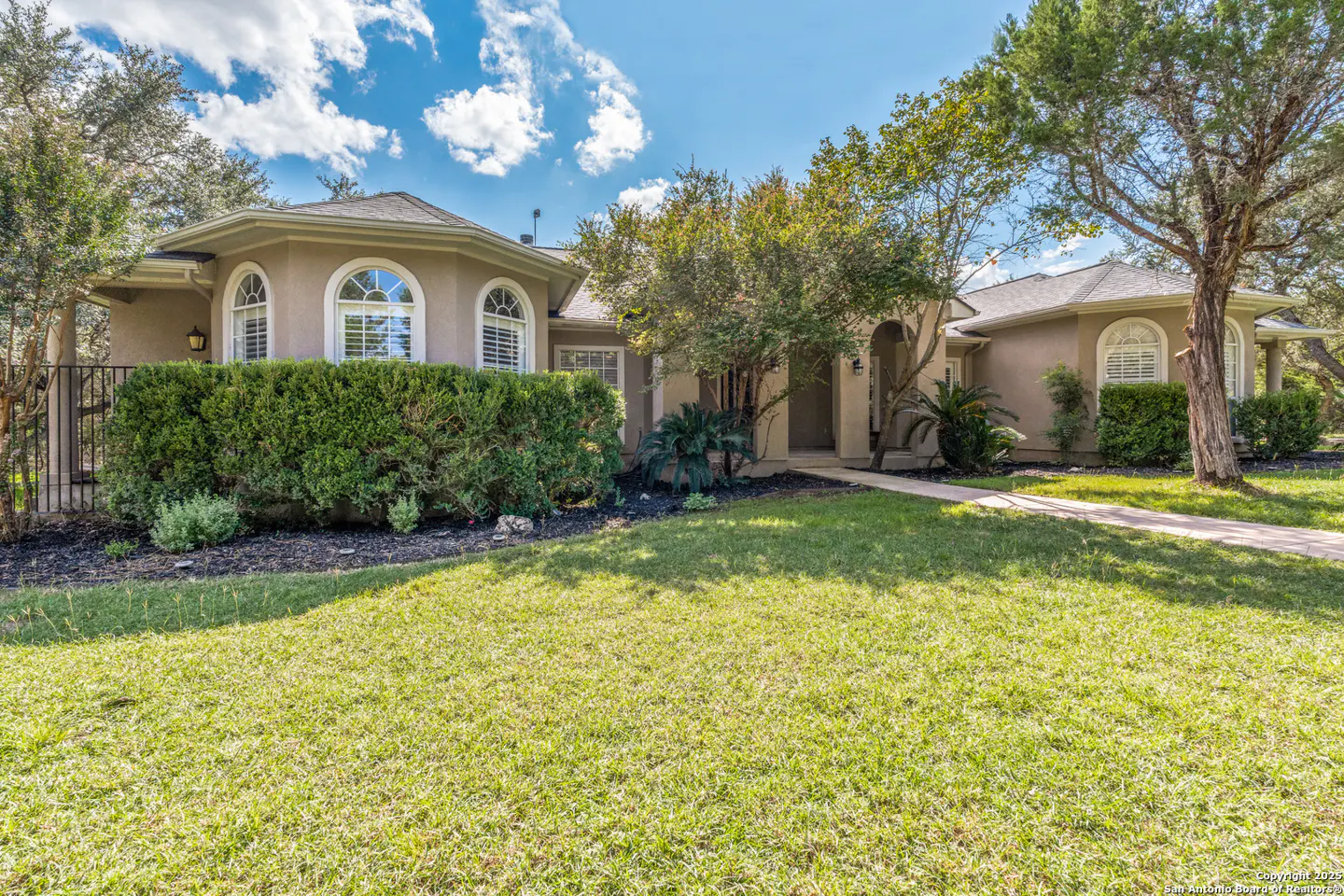 Beige single-story house with arched windows, green lawn, and trimmed hedges under a blue sky with scattered clouds.