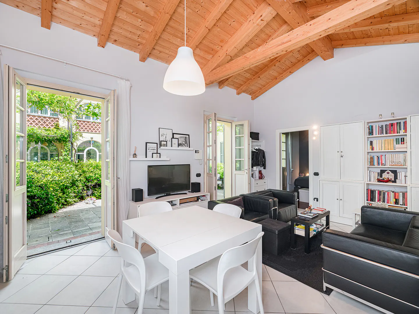 Bright living room with white walls, wood ceiling, black leather sofas, and a white table. Open doors lead to a green courtyard.