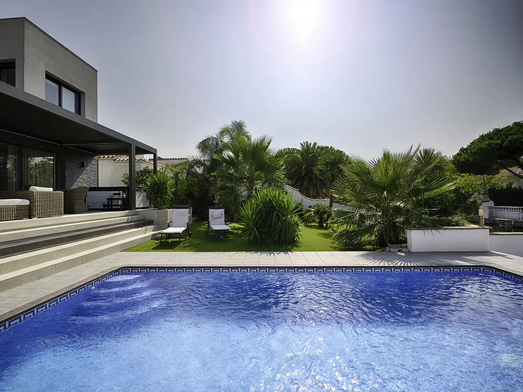 A modern home with a blue tiled pool, patio furniture, and green landscaping on a sunny day.