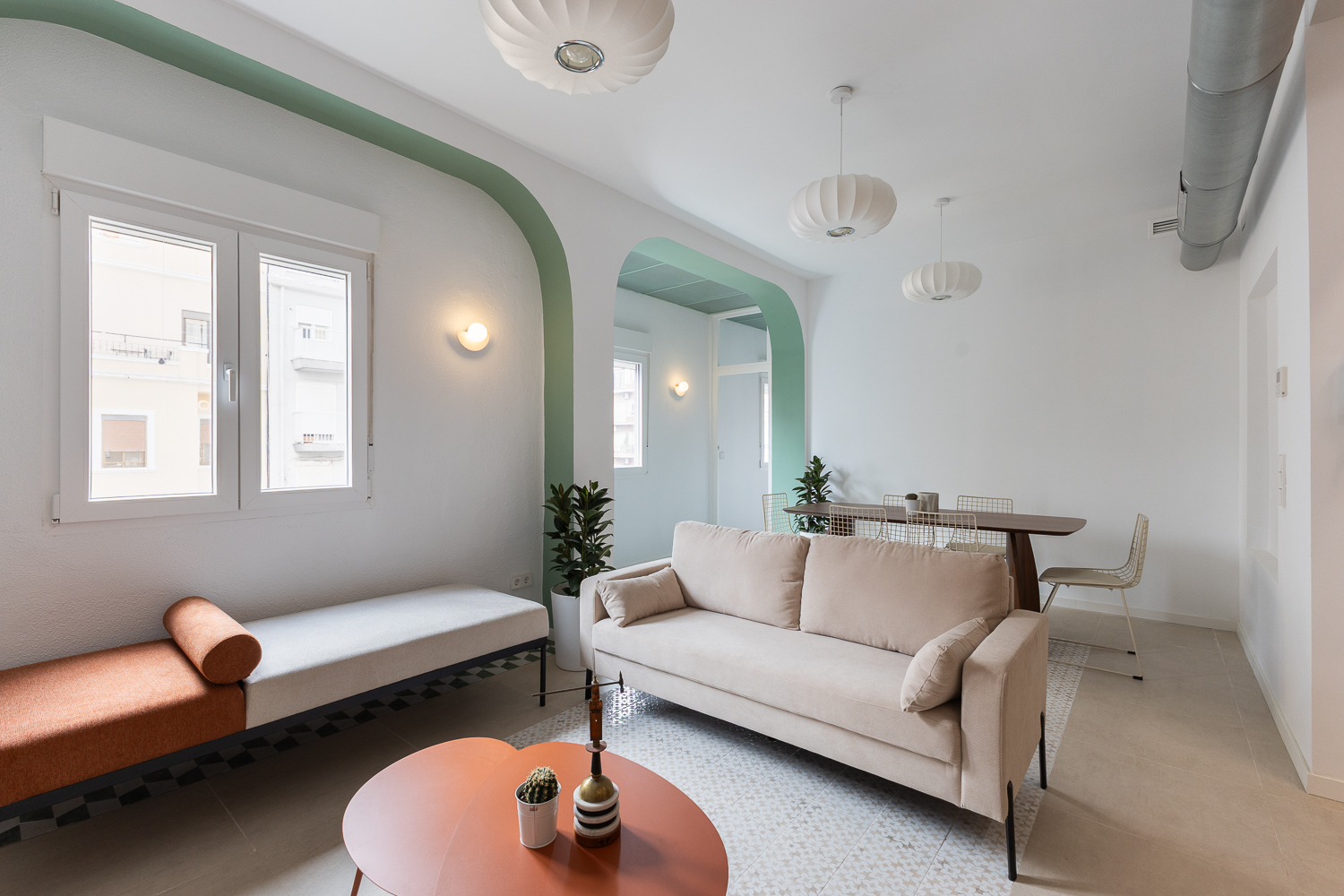 Modern living room with a beige sofa, orange daybed, round coffee table, and dining area under sage green arched walls.