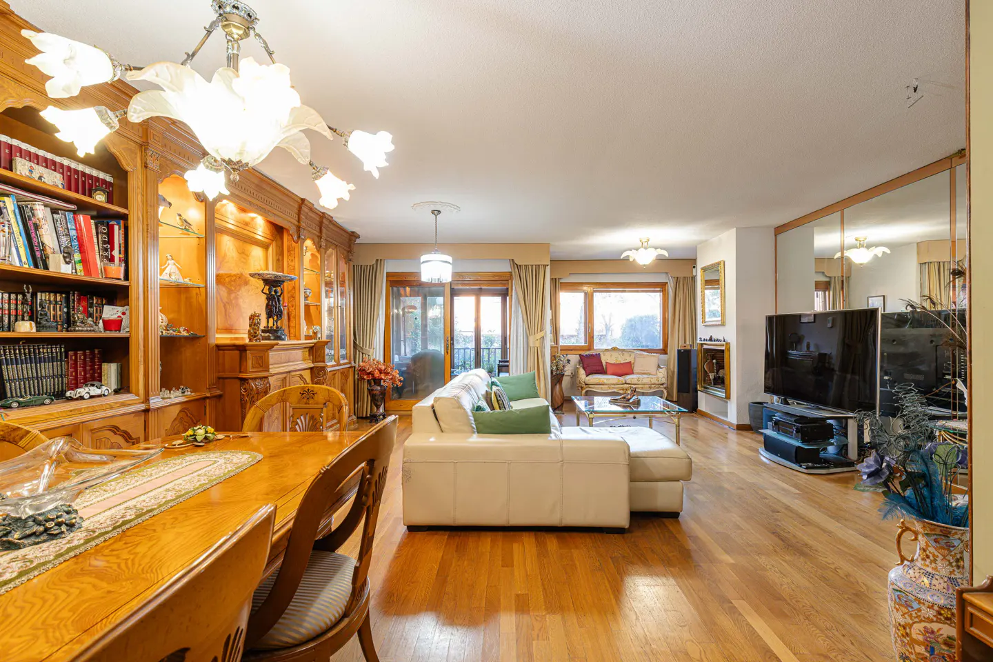 A spacious living room with hardwood floors, a white sofa, a wooden dining table, and a large bookcase.