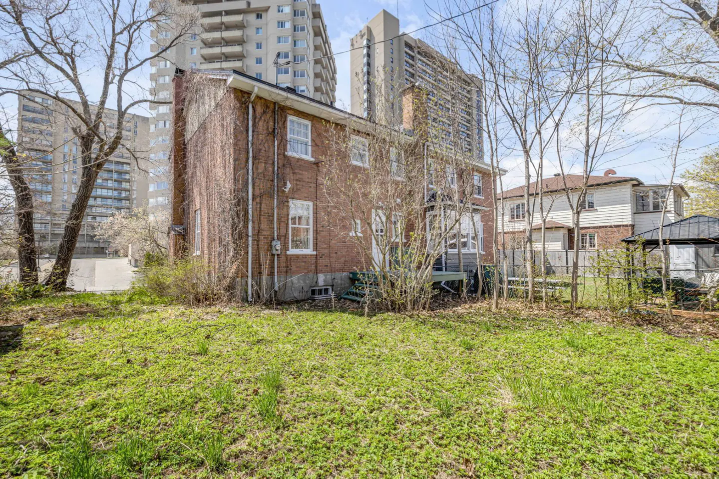 Backyard view of a two-story brick house with white windows, green lawn, and tall apartment buildings in the background.