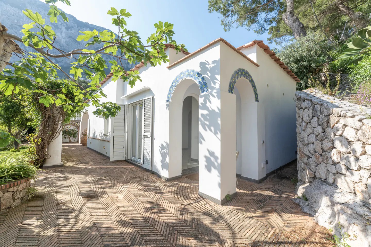 Exterior view of a white building with arched doorways and blue tile accents, surrounded by lush greenery and a stone wall.