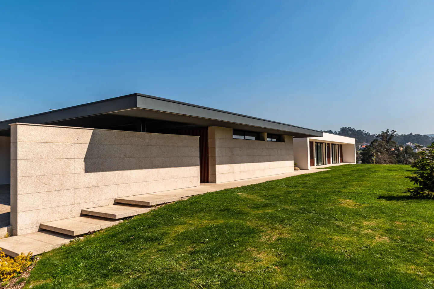 Modern single-story home with a flat roof, stone walls, and a green lawn under a clear blue sky.