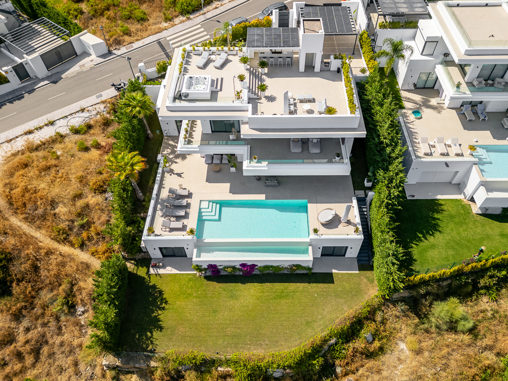 Aerial view of a modern white villa with a rooftop terrace, infinity pool, and lush green lawn in a sunny coastal area.