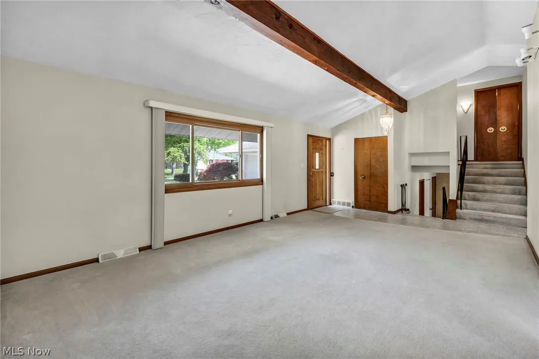 A bright, empty living room with beige carpet, a window, and a wooden beam on the ceiling. Stairs lead to a second floor.