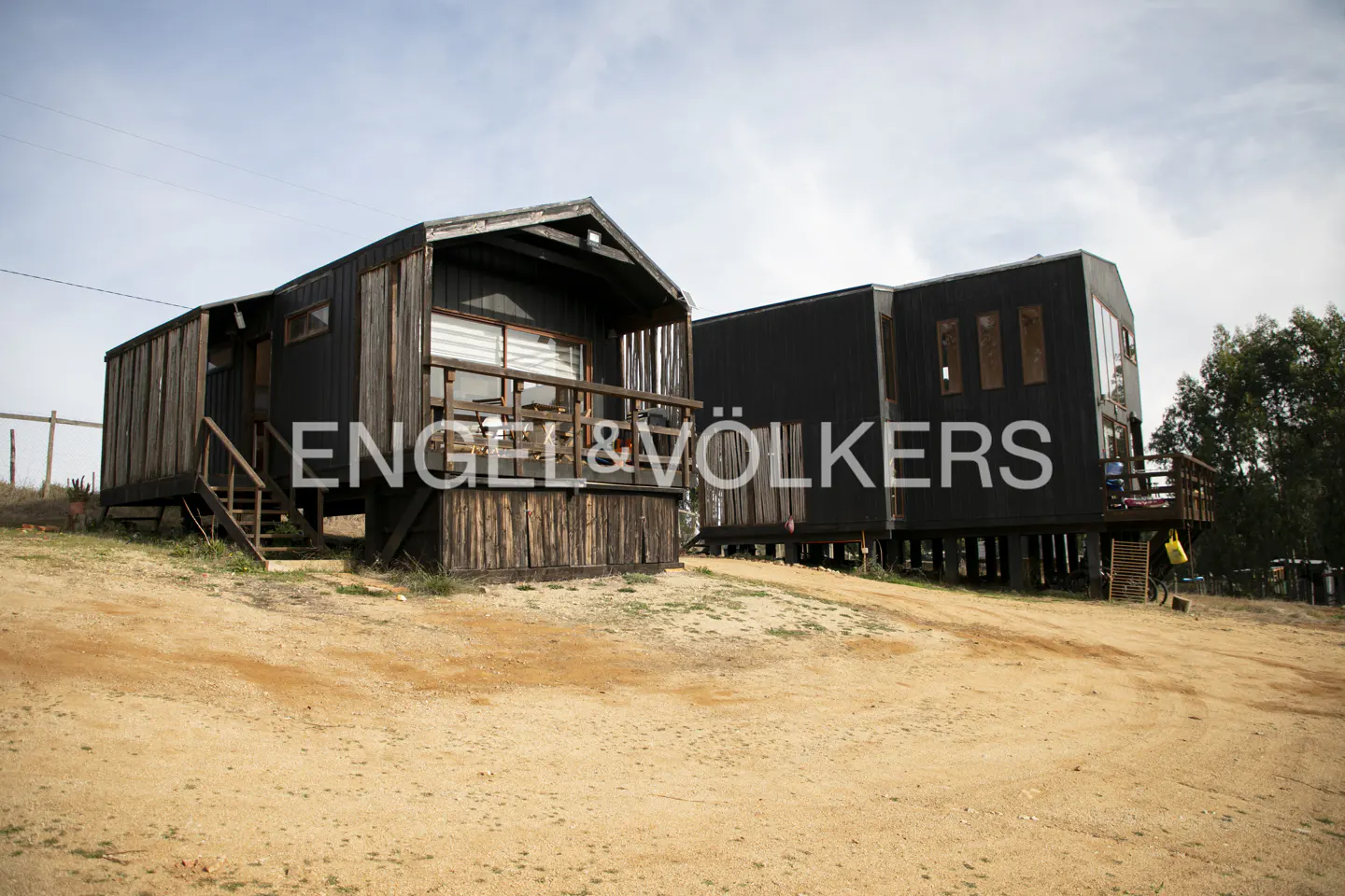 Two modern, dark-wood cabins on stilts sit on a sandy, sloped lot under a cloudy sky.