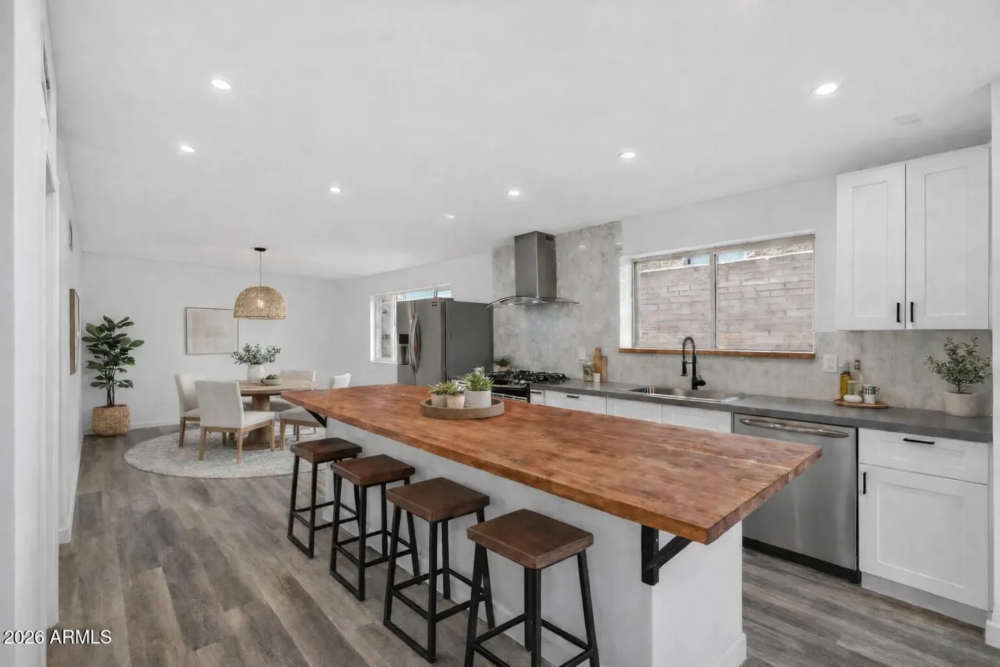 Bright, open kitchen with a wood-topped island and four stools. White cabinets and stainless steel appliances. Dining area in the background.