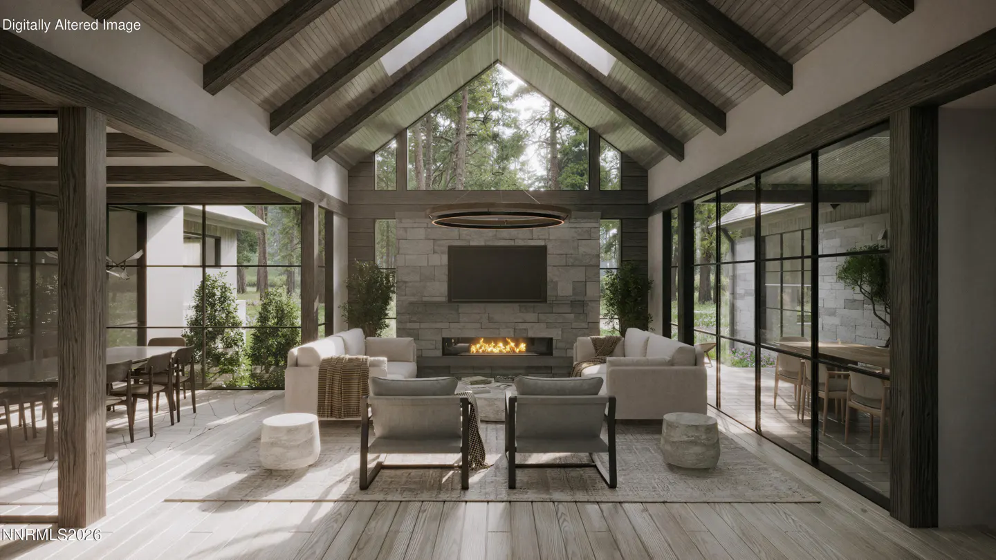 A bright living room with a stone fireplace, TV, and two sofas. Two chairs face the fireplace. Large windows offer views of trees.