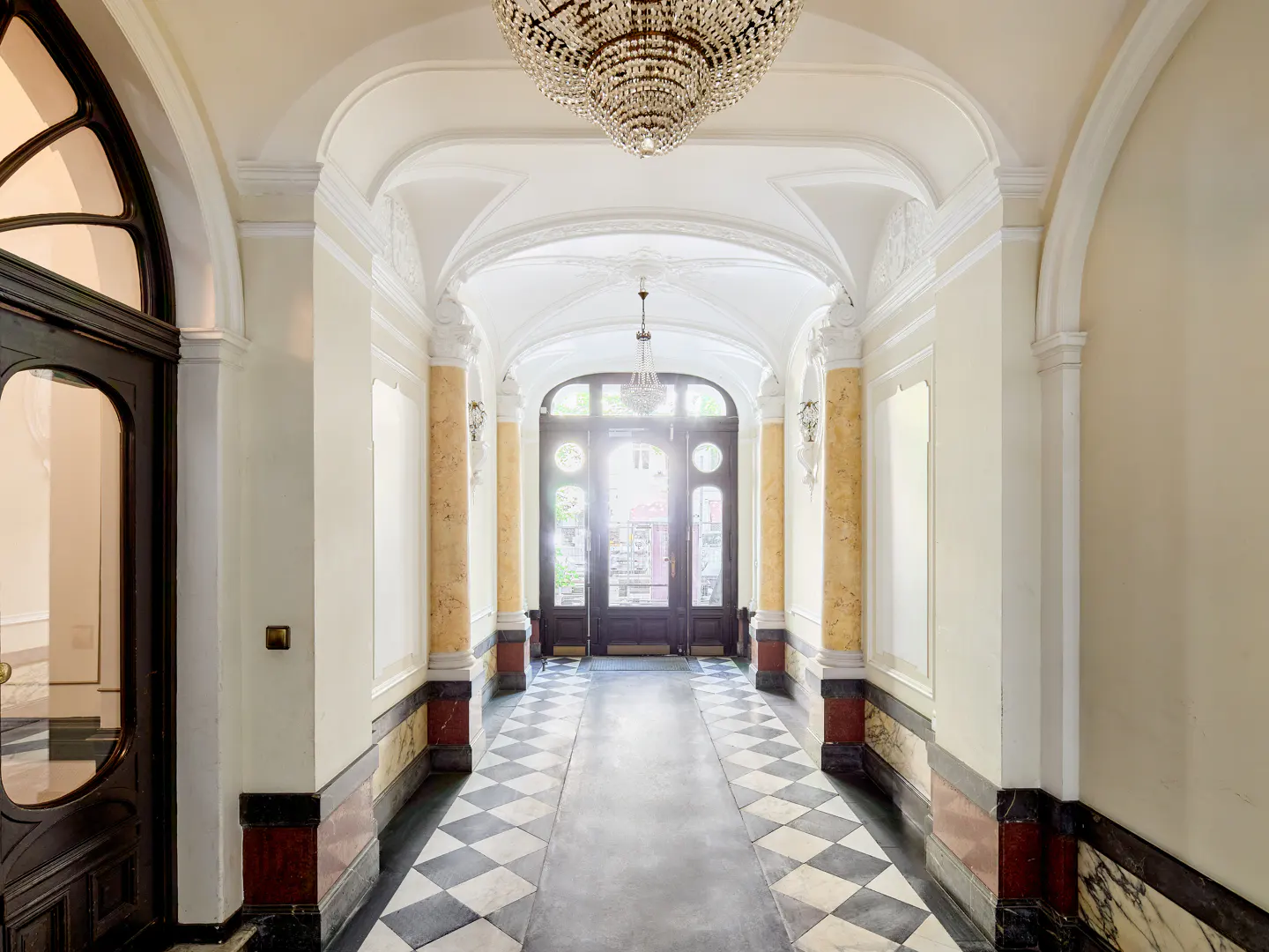 Elegant hallway with arched ceiling, crystal chandelier, and black and white checkered floor. Light shines through the front door.