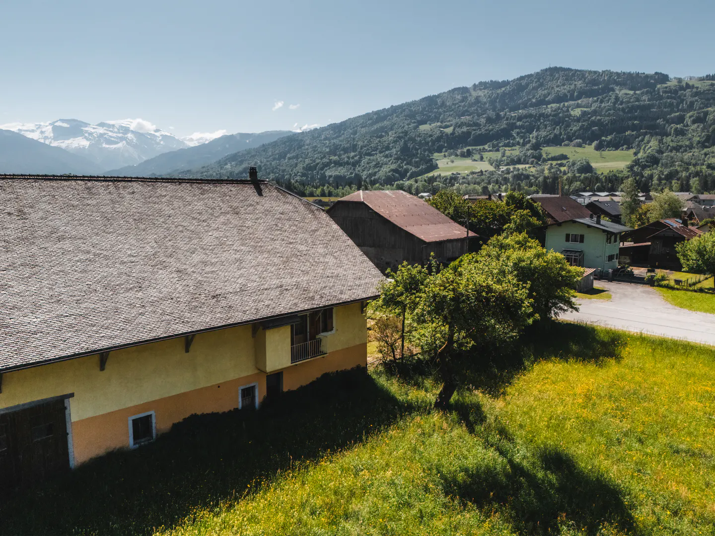 Scenic view of a yellow farmhouse with a gray roof, set against a backdrop of green hills and snow-capped mountains under a clear blue sky.