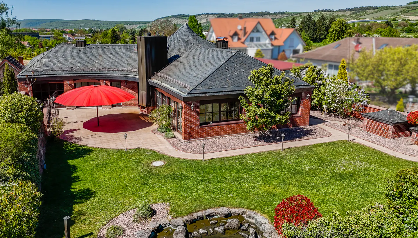 Aerial view of a red brick house with a gray roof, green lawn, red umbrella, and a small pond.