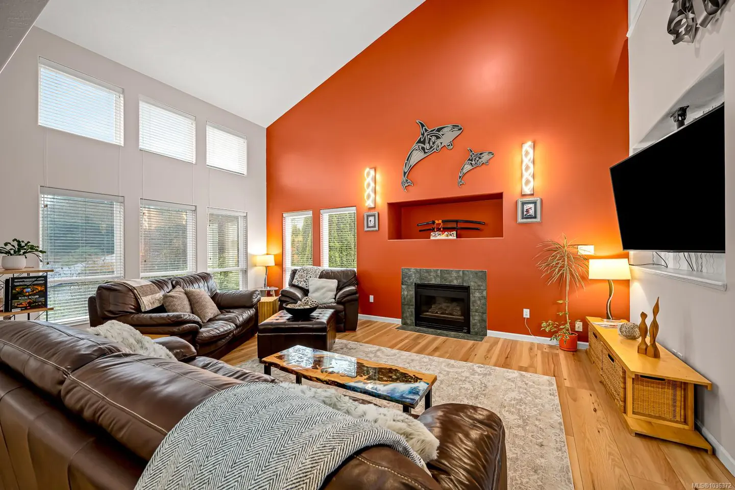 A living room with brown leather sofas, a fireplace, and an orange accent wall with orca art. Large windows provide natural light.